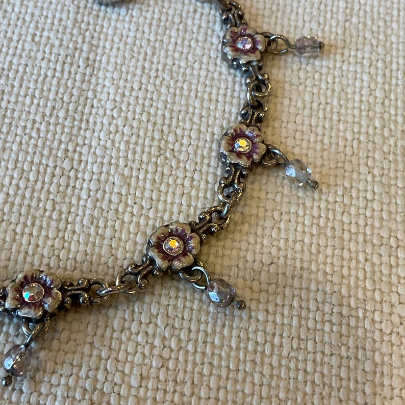 Peut inclure: Un collier &agrave; cha&icirc;ne en m&eacute;tal argent&eacute; compos&eacute; de maillons en forme de fleurs avec des d&eacute;tails en &eacute;mail violet et un strass transparent au centre. De petites perles transparentes &agrave; facettes sont suspendues entre les segments floraux.