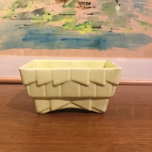 May include: A pale yellow ceramic planter with a geometric design. The planter has a rectangular shape with a slightly flared rim and four feet.