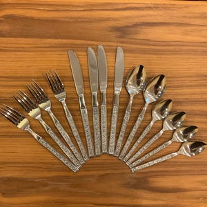 Puede incluir: Un juego de cubiertos de acero inoxidable dispuestos en abanico, que incluye cuatro tenedores, cuatro cuchillos y cinco cucharas. Cada pieza tiene un acabado de metal pulido y un mango texturizado con un diseño floral o de enredadera.