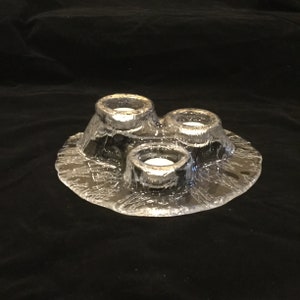 May include: A clear glass candle holder with three individual candle cups. The holder has a textured, frosted surface and a rounded, organic shape.