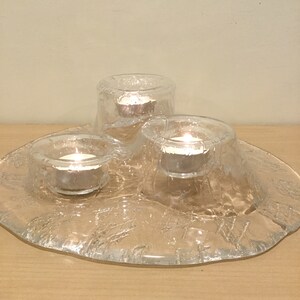 May include: A clear glass candle holder with three individual candle cups. The holder has a textured, frosted surface and a rounded, organic shape. The candle cups are filled with small, white candles.
