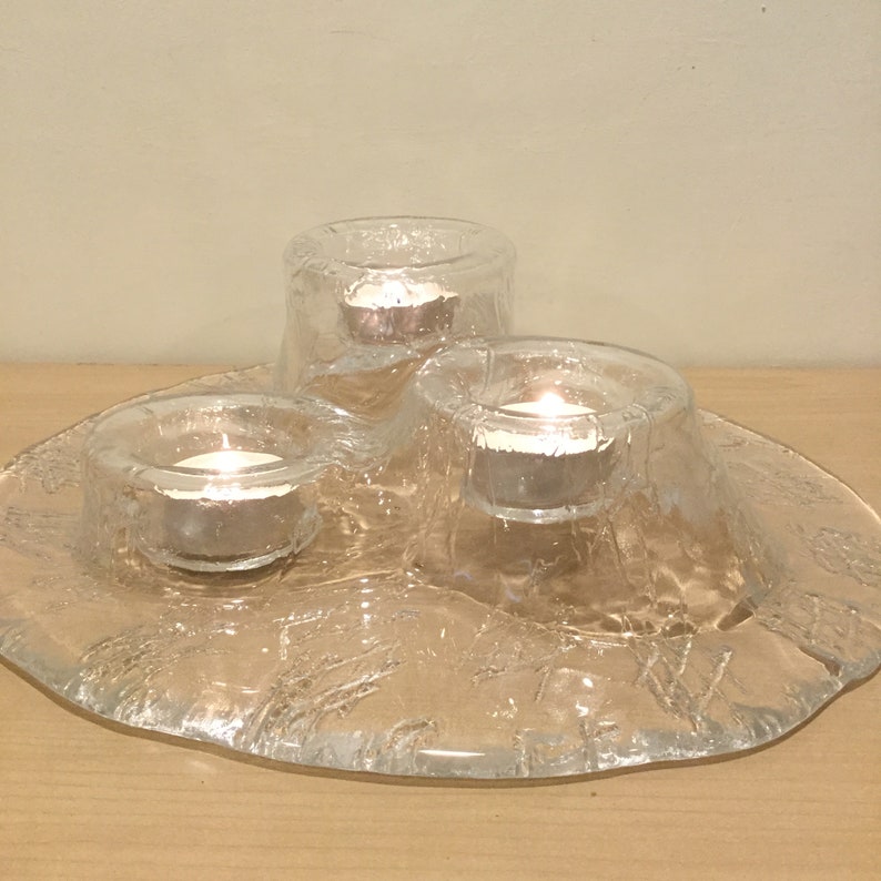May include: A clear glass candle holder with three individual candle cups. The holder has a textured, frosted surface and a rounded, organic shape. The candle cups are filled with small, white candles.