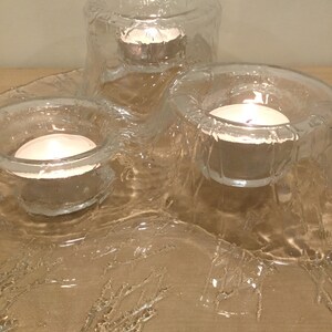 May include: A clear glass candle holder with three individual candle wells. The holder has a textured, wavy design and is lit with three tea lights.