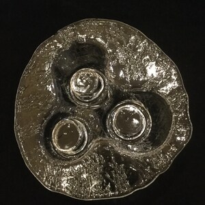 May include: A clear glass dish with three indented circular areas for holding small items. The dish has a textured, frosted surface.
