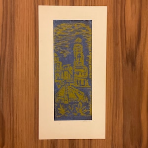 Puede incluir: Un grabado en linóleo azul y amarillo que representa una escena arquitectónica estilizada. El grabado presenta una pirámide, edificios y follaje, todo ello en un estilo gráfico audaz. El grabado está sobre un papel rectangular.