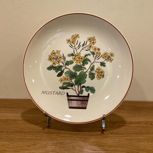 以下が含まれることがあります： 茶色の縁とマスタード植物のイラストが描かれた白いセラミックプレート。植物は、木製のポットに黄色い花と緑の葉があります。「MUSTARD」の文字が下に印刷されています。