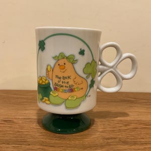 May include: White ceramic mug with a green pedestal base and a clover-shaped handle. The mug features a cartoon leprechaun with the text "The Luck o' the Irish to all Mums & Dads" and a pot of gold. Green shamrocks decorate the mug.