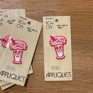 May include: A set of four pink and white iron-on patches featuring a cartoon milkshake with a cherry on top. The patches are packaged on brown cardstock with the text "Wrights Sew On 193 145 0 90¢" and "Appliques".