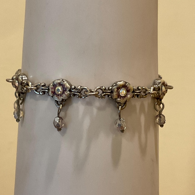 Peut inclure: Un bracelet en m&eacute;tal argent&eacute; compos&eacute; d'une s&eacute;rie de maillons en forme de fleurs, chacun orn&eacute; d'un petit cristal iris&eacute; en son centre. Le bracelet comporte des perles de cristal transparent facett&eacute;es suspendues aux fleurs par des anneaux.