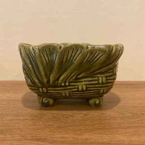 May include: A green ceramic planter with a textured, leaf-like design around the rim and a woven basket pattern on the sides. The planter is supported by three small, rounded feet. The overall shape is rectangular.