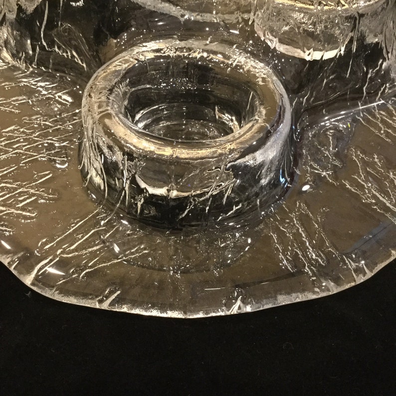 May include: A clear glass candle holder with a textured surface and a round, raised center. The holder has four indentations for candles.