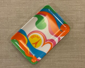 May include: A colorful, rectangular tray with a swirl pattern in shades of blue, green, orange, and pink. The tray has a white background and features a logo with the text "BEHM" and a stylized Eiffel Tower.