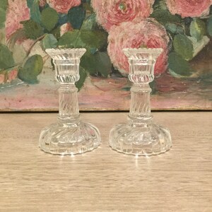 May include: Two clear glass candle holders with a spiral design on the stem and base. The candle holders have a scalloped edge at the top. The background features a painting of pink roses.