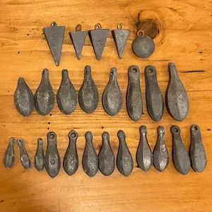 May include: A selection of grey fishing weights in various shapes, including teardrops, triangles, and spheres. The metal weights have loops for attaching to fishing line. They are displayed on a wooden surface.