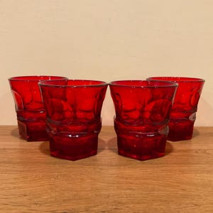 May include: Four red glass tumblers with a textured design. The glasses have a geometric shape and are arranged on a wooden surface. The tumblers are a deep red color and appear to be made of glass.