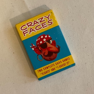 May include: A blue and yellow box with the words "Crazy Faces" printed on the front. The box features a cartoon pirate with a red bandana and an eyepatch. The text "Two Complete Card Games "Eights" and "I Doubt It" is printed on the bottom of the box.