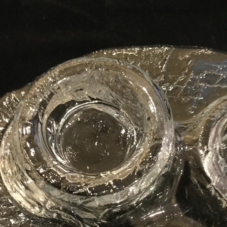 May include: A clear glass candle holder with three circular depressions for candles. The glass has a textured surface.