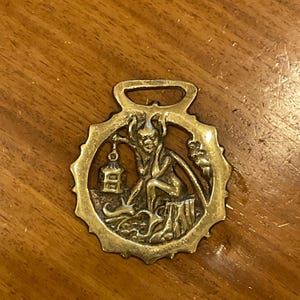 May include: A brass horse brass featuring a devil-like figure seated on a tree stump, holding a lantern. The figure has horns and a tail. The brass has a decorative border and a loop at the top for hanging.