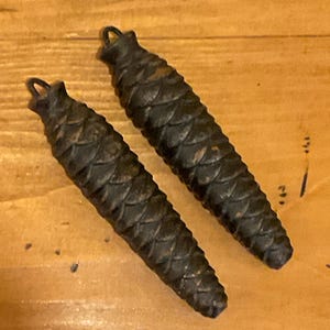May include: Two dark brown, cone-shaped clock weights with a textured, scaled surface. Each weight has a small loop at the top. The weights are lying on a light brown wooden surface.