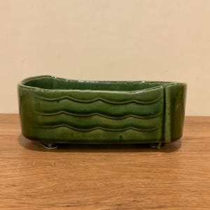 May include: A rectangular ceramic planter with a glossy dark green glaze. The exterior features a horizontal wavy relief pattern along the sides. The vessel is supported by four small feet at the base and has an open top with a smooth rim.