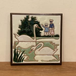 May include: A square ceramic tile framed in dark wood featuring a relief design of two white swans swimming in a light green pond. The scene includes dark green foliage, tall reeds, and three small figures of children standing on the grassy bank.