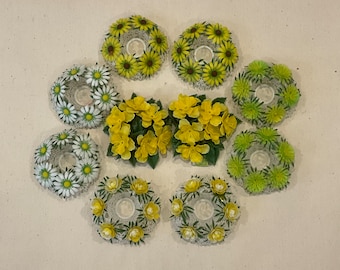 May include: Nine white and yellow floral wreaths with a center hole for a candle or tea light. The wreaths are arranged in a circle on a beige surface.