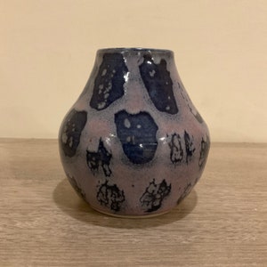 May include: A small, round ceramic vase with a light purple and dark blue speckled glaze. The vase has a wide base and a narrow neck.