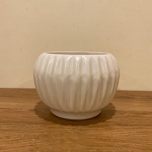 May include: A round, off-white ceramic planter featuring a distinct vertical ribbed texture across its exterior surface. The vessel has a wide, open top and a smooth, rounded body that tapers slightly toward a flat, circular base at the bottom.