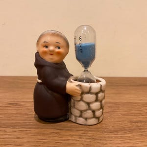 May include: A ceramic figurine of a monk in a brown habit standing beside a white stone-textured well. The monk holds a glass hourglass containing blue sand, with black numerical markings 3, 4, and 5 printed vertically on the glass surface.