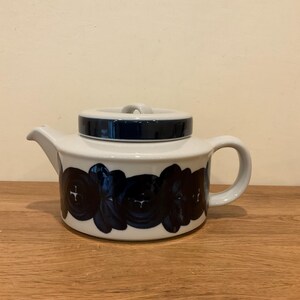 May include: A white ceramic teapot featuring a dark blue floral pattern around the base. The teapot has a matching lid with a dark blue band, a curved handle on the right side, and a short spout on the left side, presenting a low-profile design.