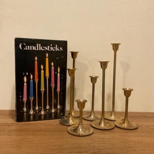 May include: A collection of brass candlesticks of varying heights, alongside a box labeled "Candlesticks" with an illustration of lit candles in different colors. The candlesticks are arranged on a wooden surface.