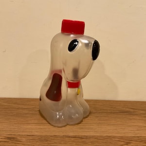 Puede incluir: Una botella de plástico transparente con forma de perro con tapa, collar y mancha marrón rojos. El perro tiene ojos negros grandes y está sentado sobre una superficie de madera. La botella es un objeto de colección vintage.
