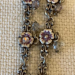 Peut inclure: Un collier &agrave; cha&icirc;ne en m&eacute;tal argent&eacute; compos&eacute; de maillons floraux r&eacute;p&eacute;t&eacute;s avec des d&eacute;tails en &eacute;mail blanc et violet. Chaque centre de fleur est orn&eacute; d'un petit cristal clair iris&eacute;, et la cha&icirc;ne est agr&eacute;ment&eacute;e de perles en verre transparent &agrave; facettes.