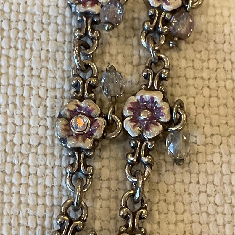 Peut inclure: Un collier &agrave; cha&icirc;ne en m&eacute;tal argent&eacute; compos&eacute; de maillons floraux r&eacute;p&eacute;t&eacute;s avec des d&eacute;tails en &eacute;mail blanc et violet. Chaque centre de fleur est orn&eacute; d'un petit cristal clair iris&eacute;, et la cha&icirc;ne est agr&eacute;ment&eacute;e de perles en verre transparent &agrave; facettes.