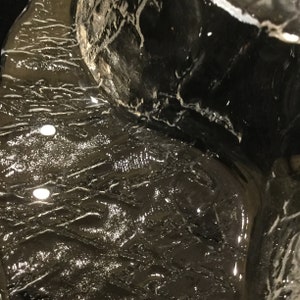 May include: Close-up of a dark gray, textured surface with a reflective, wet appearance. The surface has a pattern of white lines and streaks, creating a unique and abstract design.