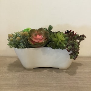 May include: A white ceramic planter filled with a variety of artificial succulents. The succulents are in shades of green, red, and pink. The planter is rectangular with rounded edges and sits on a wooden surface.