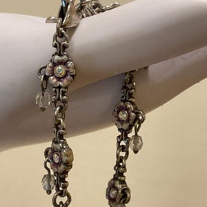 Peut inclure: Un bracelet &agrave; cha&icirc;ne en m&eacute;tal argent&eacute; compos&eacute; de maillons en forme de fleurs avec des accents en &eacute;mail violet et des centres en cristal transparent &agrave; facettes. Chaque maillon floral est reli&eacute; par des anneaux en m&eacute;tal et inclut une perle.