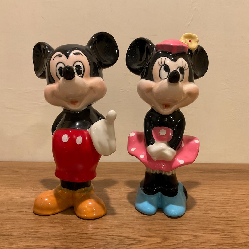 Op de afbeelding: Een vintage set keramische beeldjes van Mickey Mouse en Minnie Mouse. Mickey draagt een rode broek met witte stippen en gele schoenen. Minnie draagt een roze jurk met witte stippen, een blauwe strik en blauwe schoenen.