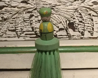 May include: A green and yellow wooden doll with a green hat and a green and white brush attached to the bottom.