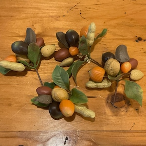 Könnte beinhalten: Eine Anordnung aus künstlichen Nüssen und Früchten, darunter Erdnüsse, Walnüsse und kleine orangefarbene Früchte, mit grünen Blättern. Die Anordnung befindet sich auf einer Holzoberfläche.