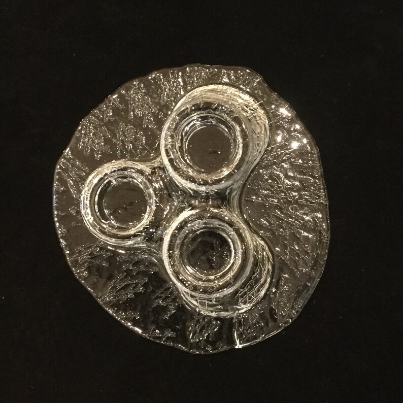 May include: A clear glass candle holder with three circular depressions for candles. The holder has a textured, frosted surface.