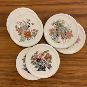 May include: A set of five round white ceramic coasters featuring colorful illustrations of birds, flowers, and a traditional carriage. Each coaster has a raised rim and displays detailed artwork in shades of orange, pink, blue, and green.
