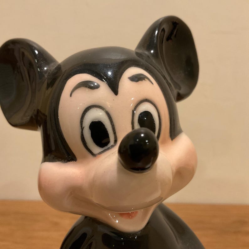 Op de afbeelding: Een keramiek beeldje van Mickey Mouse. Het beeldje is zwart en wit met een zwarte neus en grote, ronde ogen. Mickey Mouse glimlacht en heeft een zwart-wit oor aan de linkerkant van de afbeelding.