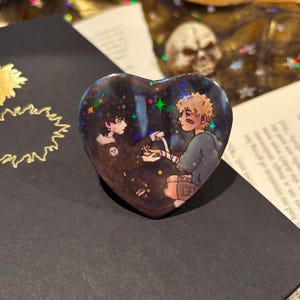 May include: Heart-shaped pin with a holographic finish, featuring an illustration of two figures. One figure is wearing a skull-themed shirt. The other figure has a bandage on their arm. The background is dark with star-like accents.