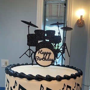 May include: A round white cake decorated with black musical notes and a black border. A black glitter drum set cake topper with the words "Happy Birthday" is on top. The cake is on a white cake board.