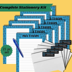 May include: A 21-piece stationery kit featuring lined paper with a blue border of pixelated handheld game consoles and the name Nathan printed at the top. The set includes white envelopes, black address labels with blue text, and a blue ballpoint pen.