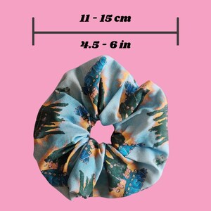 May include: A blue scrunchie with a repeating pattern of buildings and trees. The scrunchie measures 11 - 15 cm in diameter. The background is a solid pink colour.
