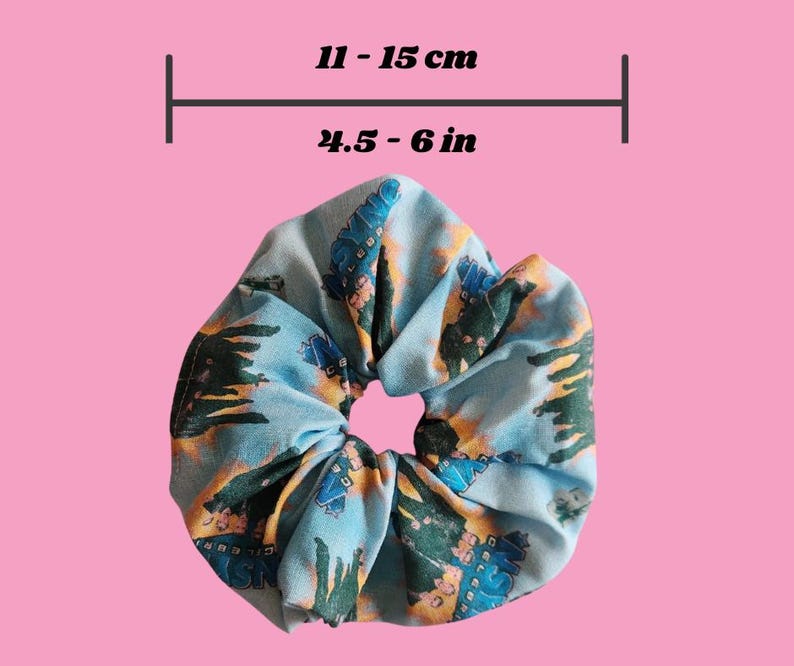 May include: A blue scrunchie with a repeating pattern of buildings and trees. The scrunchie measures 11 - 15 cm in diameter. The background is a solid pink colour.