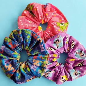 May include: Three colorful scrunchies on a blue background. One is pink with a cartoon character print, another is blue with a cartoon print, and the third is purple with a cartoon print. These fabric hair accessories are a fun and stylish way to accessorize.