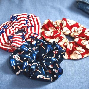 May include: Three fabric scrunchies with patriotic and sports-themed patterns. One features the American flag, another baseballs on red, and the third has blue popsicles and stars on a navy background. These hair accessories are perfect for adding a touch of flair.